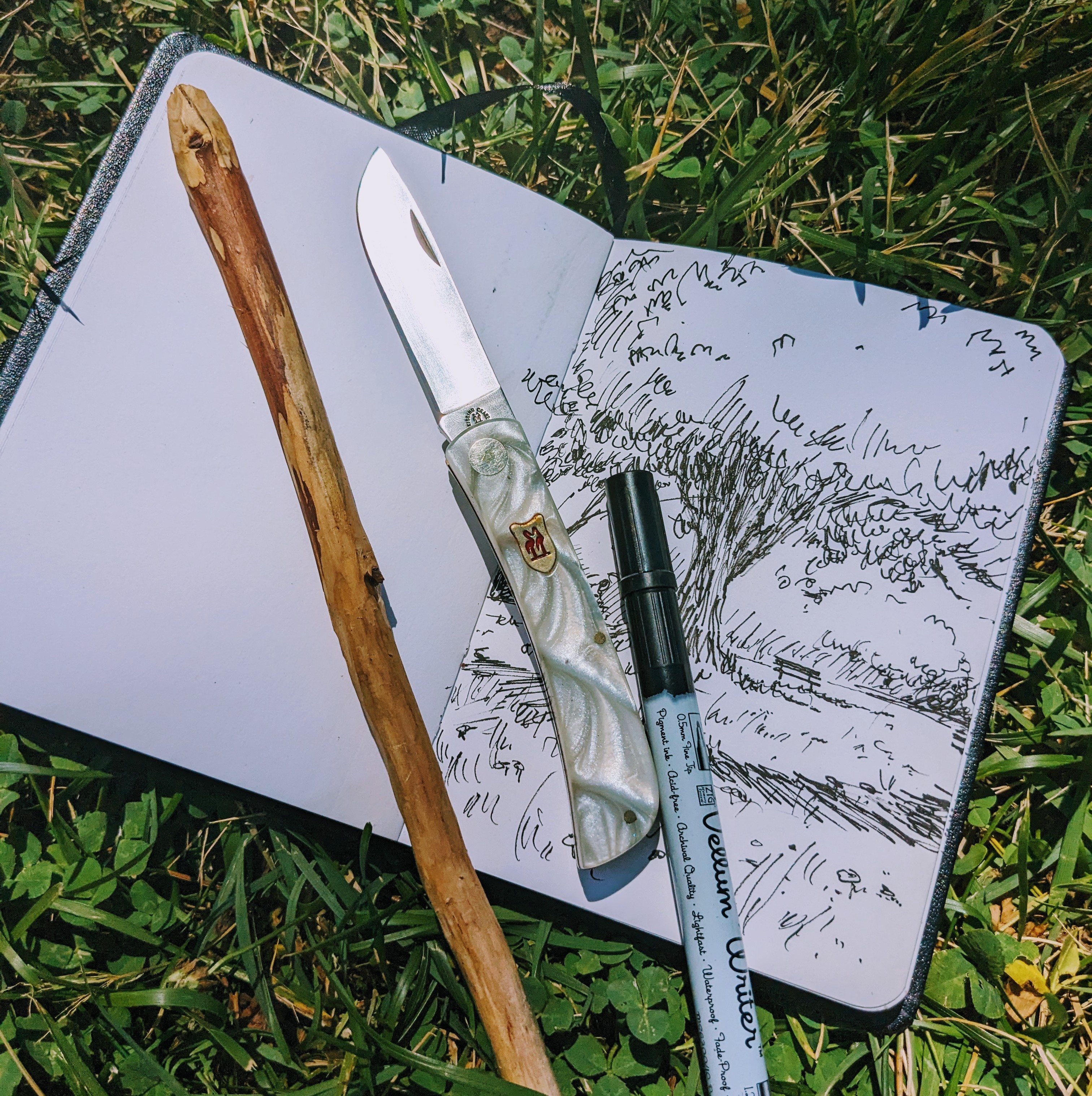 A photo of a notebook with a drawing, a pocket knife, and a stick to whittle on